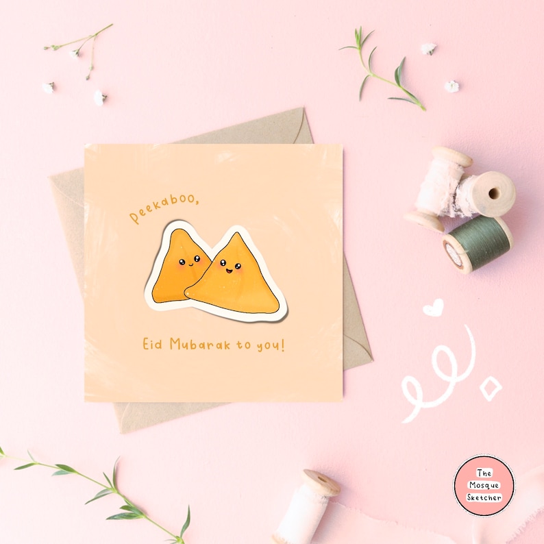 May include: A peach-colored card with a cartoon illustration of two samosas with smiling faces. The text "Peekaboo, Eid Mubarak to you!" is written in brown lettering.