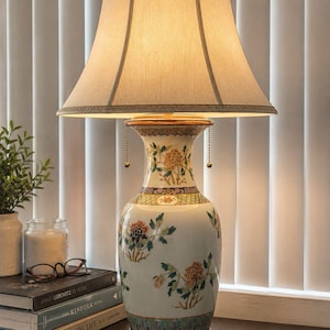 May include: A table lamp with a beige fabric bell-shaped shade and a ceramic base featuring a white background with hand-painted floral motifs in orange, green, and blue. The base has a dark wooden stand and two gold-toned pull chains for operation.
