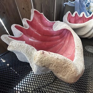 Puede incluir: Dos grandes cuencos decorativos con forma de concha, con un exterior texturizado en color blanco roto y un interior liso, brillante y de color rosa intenso. Las conchas presentan un diseño distintivo de bordes ondulados y festoneados.
