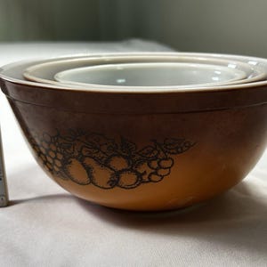 May include: A set of two nested mixing bowls with a brown gradient exterior and a dark brown fruit illustration. A vertical measuring tape is placed next to the bowls, showing the height of the larger bowl is approximately 3.5 inches.