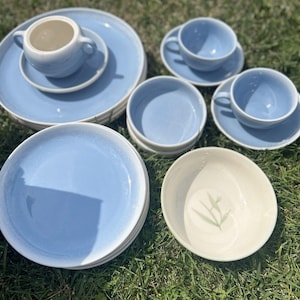 May include: A ceramic dinnerware set featuring light blue plates, two teacups with matching saucers, a small lidded sugar bowl, and a cream-colored bowl with a green leaf illustration. The collection includes multiple stacked plates and bowls.