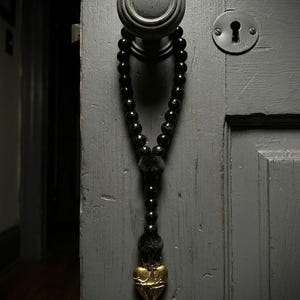 May include: A string of glossy black beads hanging from a round gray door knob. The beads are separated by small black fuzzy pom-poms and terminate in a gold-colored metal heart pendant featuring an embossed design of a crown and thorns.
