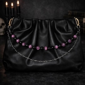 May include: A black gathered handbag featuring a decorative strap composed of alternating purple and black beads, complemented by a thin dark metal chain. The bag is secured with gold-toned metal clasps on each side of the top opening.