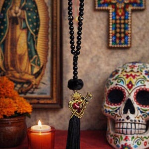 Puede incluir: Un adorno colgante que presenta una hilera de cuentas negras, un pequeño pompón negro y un dije de metal dorado con forma de corazón atravesado por una espada. El corazón tiene un centro rojo y el adorno termina con una borla de tela negra.