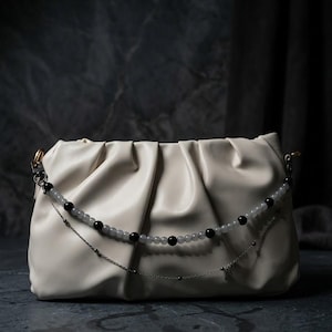 May include: A cream-colored, pleated clutch bag featuring a decorative double-strand accessory. The top strand consists of alternating black and translucent grey beads, while the bottom strand is a thin silver-toned metal chain with small accents.