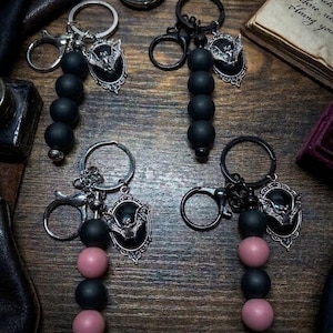 May include: Four keychains featuring silver-toned bat charms set in black oval frames. Two keychains have four matte black beads, while two feature alternating matte black and dusty pink beads. Each includes a metal split ring and a lobster clasp.