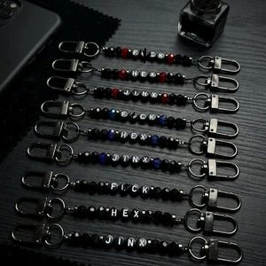 May include: A collection of nine beaded lanyards featuring black metal lobster clasps at both ends. Each lanyard contains black beads, some with red or blue accents, and letter beads spelling out FUCK, HEX, or JINX in a repeating sequence.