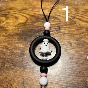May include: A black and white Halloween-themed car charm. The charm features a white ghost inside a black ring, with black, pink, and white beads, and a black tassel. The number "1" is visible in the background.
