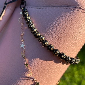 May include: Black beaded charm with gold star accents, attached to a pink bag. The charm features a chain of gold stars and crescent moons. The black beads have gold star patterns. The charm is attached to a black clasp.