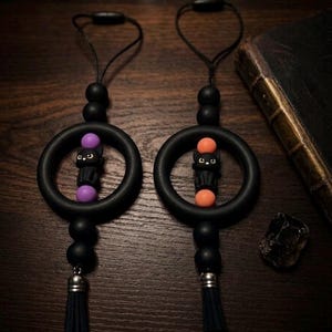 May include: Two black circular charms featuring a central black cat figure flanked by either purple or orange beads. Each charm includes a black cord loop, additional matte black beads, and a small silver-toned metal cap above a black tassel.