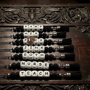 May include: A collection of beaded necklaces with black tassels and silver-toned accents. Each necklace features white lettered cubes spelling out words like "VAMP," "HEX," "JINX," "GOTH," "NURSE," and "TEACH." The background includes carved wooden details and a skull.