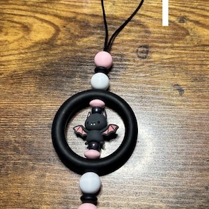 Puede incluir: Un dije negro con forma de murciélago con alas y detalles en rosa, centrado dentro de un anillo negro. El dije está suspendido de un cordón negro con cuentas rosas y blancas. El fondo es una superficie de madera.