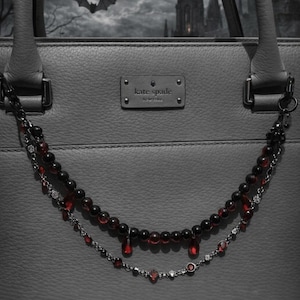 May include: Gray Kate Spade handbag with a decorative chain featuring black beads and red teardrop-shaped accents. The background shows a spooky scene with a bat, a castle, and a cloudy sky.