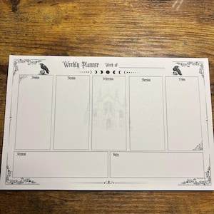 May include: A white weekly planner with a gothic theme. The planner features a raven illustration in each corner, with the words "Weekly Planner" at the top. The days of the week are listed, with space for notes and a central illustration of a gothic building.