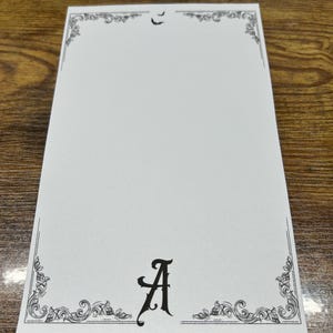 May include: A white notepad with a decorative black border and the letter "A" in a stylized font. The top of the notepad features a small bat silhouette. The design has a gothic or vintage aesthetic.