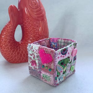 May include: A bright orange ceramic fish-shaped pitcher stands behind a small, square fabric basket. The basket is constructed from patchwork fabric in shades of pink and white, featuring a felt heart, green elephant print, and small buttons.
