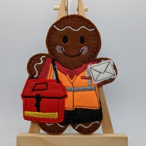 May include: A felt gingerbread man ornament depicting a postman. The brown figure wears an orange vest and carries a red mail bag and a letter. The ornament is displayed on a small wooden easel.