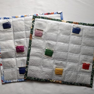 May include: Two white fabric storage organisers with clear plastic pockets for holding embroidery floss. The organiser on the left has a blue and white patterned fabric with a snowman design. The organiser on the right has a green and white patterned fabric with a floral design. Both organisers have a variety of coloured embroidery floss in the pockets.
