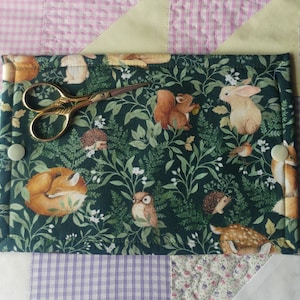 May include: A green fabric pouch with a woodland animal print. The pouch has a button closure and is decorated with a variety of animals, including foxes, squirrels, rabbits, and deer. The pouch is ideal for storing small items, such as needles, thread, or buttons.