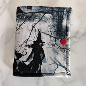 May include: A black and white fabric book cover featuring a silhouette of a witch and a cat in a woodland scene. The cover has a red button fastening.