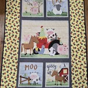 Op de afbeelding: Een handgemaakte quilt met boerderijdieren en tekst. De quilt heeft een gele rand met een tractormotief. De middelste panelen tonen varkens, schapen, koeien en paarden met zinnen als "Oink", "Baa", "Moo" en "giddy up".