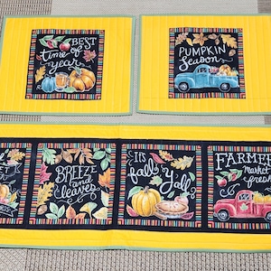 Puede incluir: Un juego de seis caminos de mesa acolchados con temática otoñal. Los caminos de mesa presentan diseños de estilo pizarra negra con motivos otoñales, incluyendo calabazas, hojas y camiones. El texto en los caminos de mesa incluye frases como "Mejor época del año", "Temporada de calabazas", "Mercado de agricultores" y "Es otoño".