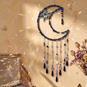 May include: A crescent moon wall hanging featuring a dark blue stone-encrusted frame, a central flower-shaped crystal accent, and multiple dangling silver chains adorned with various colourful beads and dark pointed crystal prisms.