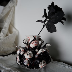 May include: A unique ceramic vase with a white base, black accents, and a design featuring multiple faces and eyes. A black rose is placed in the vase. The vase has a gothic or horror theme.
