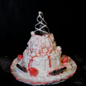 Puede incluir: Un pastel de cerámica blanco de dos niveles con detalles en rojo y una parte superior negra en espiral. El pastel está decorado con flores rojas, objetos blancos redondos y goteos rojos. El pastel está sobre un plato blanco con borde rojo.