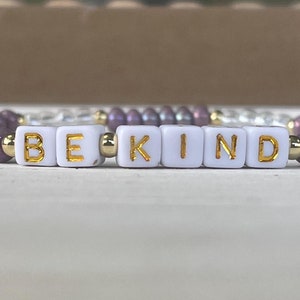 May include: A purple and white beaded bracelet with gold letter beads spelling out "BE KIND".