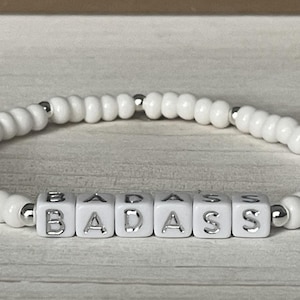May include: White beaded bracelet with silver beads and white letter beads spelling out "BADASS".