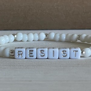 May include: White beaded bracelet with the word "RESIST" spelled out in white letter beads.