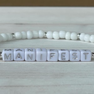 May include: White beaded bracelet with the word "MANIFEST" spelled out in silver lettering on white beads.