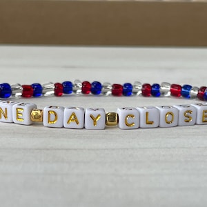 May include: A white beaded bracelet with gold lettering spelling "ONE DAY CLOSER" and red and blue beads.