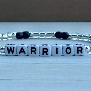 May include: A beaded bracelet with the word "WARRIOR" spelled out in white letter beads. The bracelet features clear and black beads, strung together to form a flexible band. The bracelet is displayed on a light blue surface.
