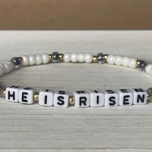 May include: White beaded bracelet with gold and silver accents spelling out "HE IS RISEN".