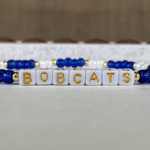 May include: A beaded bracelet with the word "BOBCATS" spelled out in gold letters on white square beads. The bracelet features alternating blue and white beads, with gold accents. This accessory is perfect for fans of the Bobcats.