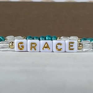 May include: A teal and white beaded bracelet with the word "GRACE" spelled out in gold letters on white beads.