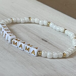 May include: White beaded bracelet with gold accents spelling out "Angel Mama".