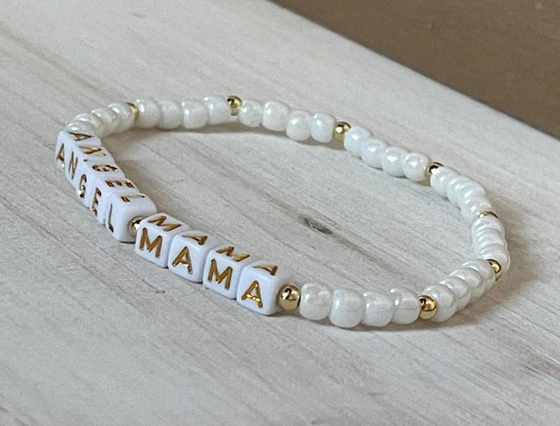 May include: White beaded bracelet with gold accents spelling out "Angel Mama".