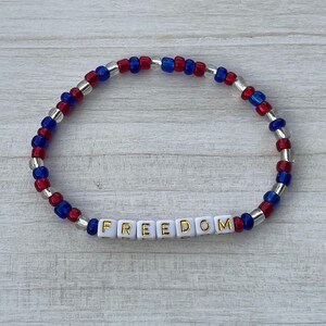 Puede incluir: Una pulsera de cuentas rojas, blancas y azules con la palabra "FREEDOM" escrita en letras doradas sobre cuentas blancas.
