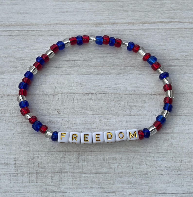 Puede incluir: Una pulsera de cuentas rojas, blancas y azules con la palabra "FREEDOM" escrita en letras doradas sobre cuentas blancas.