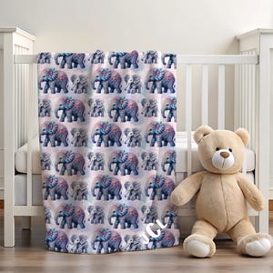 May include: A baby blanket with a repeating pattern of blue and pink elephants. The blanket is draped over a white crib, next to a beige teddy bear. The blanket has the initials "JCL" in the bottom right corner.