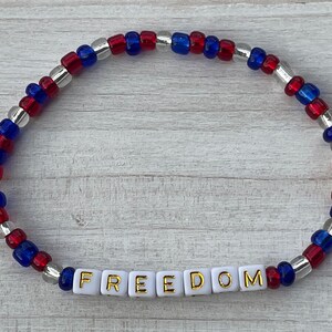 The image shows a stackable beaded bracelet with red, blue, and clear beads. It features the word FREEDOM. The bracelet symbolizes liberty, ideal for patriotic events or everyday wear. Perfect for family of military members. Freedom is not free.