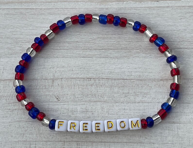 The image shows a stackable beaded bracelet with red, blue, and clear beads. It features the word FREEDOM. The bracelet symbolizes liberty, ideal for patriotic events or everyday wear. Perfect for family of military members. Freedom is not free.