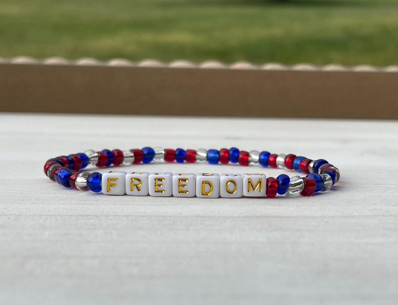 Puede incluir: Una pulsera de cuentas rojas, blancas y azules con la palabra "FREEDOM" escrita en letras doradas sobre cuentas blancas.