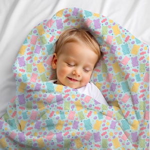 May include: A baby wrapped in a light blue blanket patterned with colorful gummy bears, hearts, stars, and candies. The blanket is soft and plush, with a pastel color scheme. The baby is sleeping peacefully.