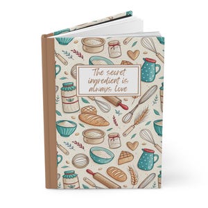 May include: A brown and cream journal with a white background and a pattern of kitchen utensils, ingredients, and wheat. The text "The secret ingredient is always love" is printed in brown on a white rectangle.