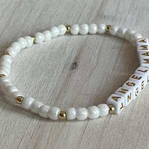 May include: White beaded bracelet with gold accents and white letter beads spelling "ANGEL MAMA".