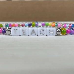 May include: A colorful beaded bracelet with white letter beads spelling out the word "TEACH".